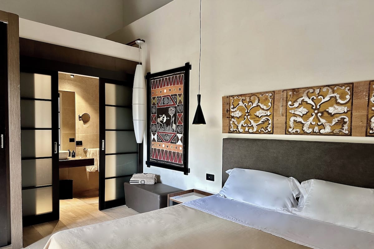 A modern hotel room with a bed, a nightstand, a mirror, a wall-mounted artwork, and a door leading to another room.