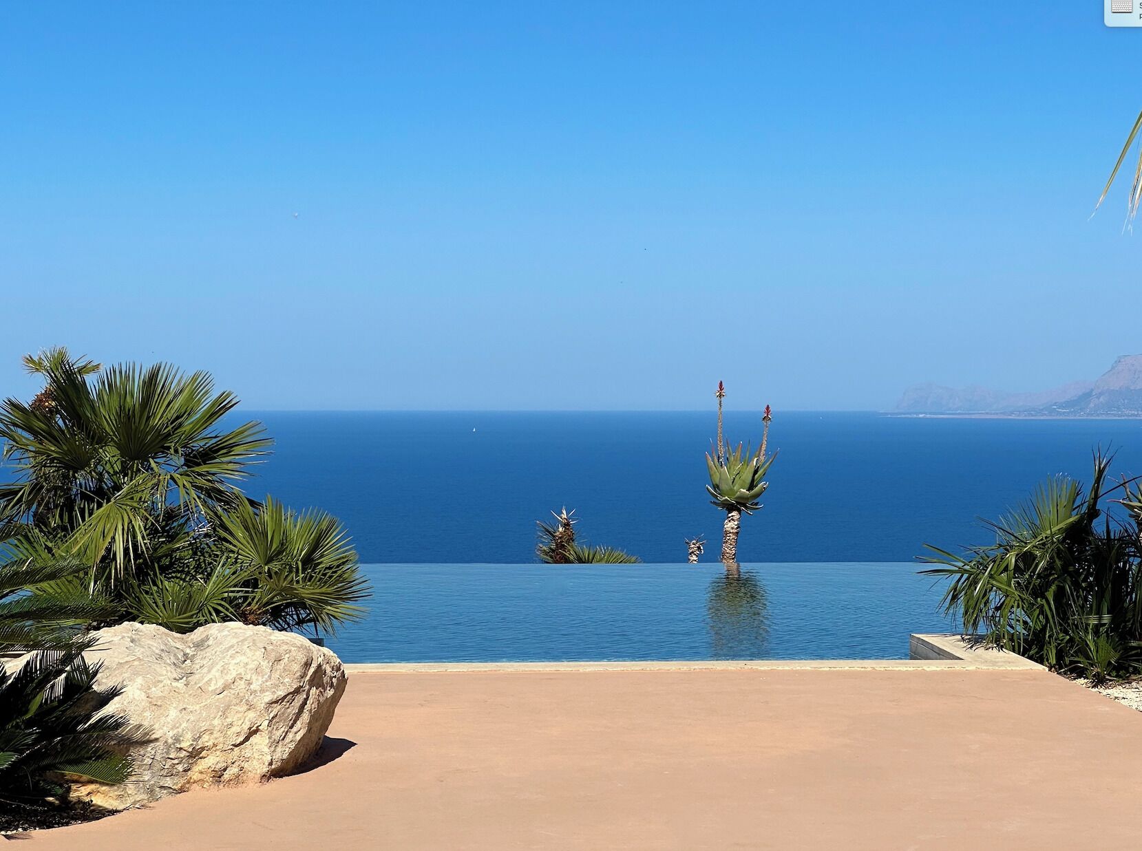 Vista despejada del océano desde la piscina infinita del resort, con palmeras y una roca grande al frente.