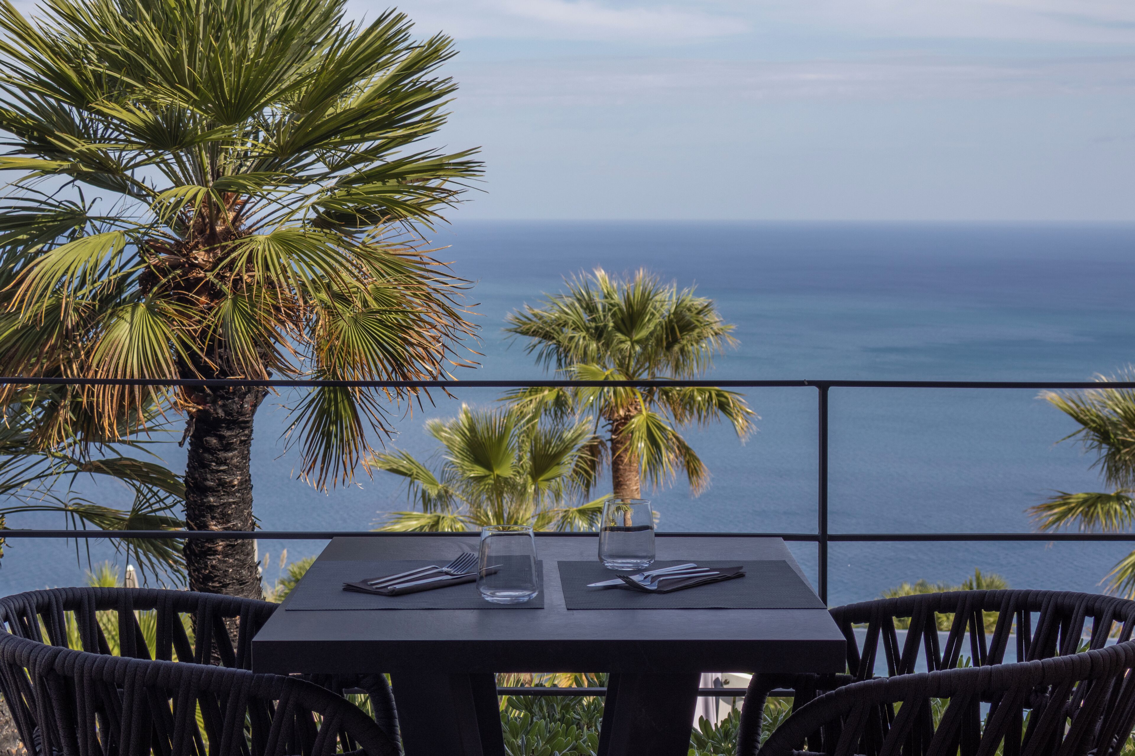 A table with two chairs, a glass, and a fork, overlooking the ocean with palm trees.