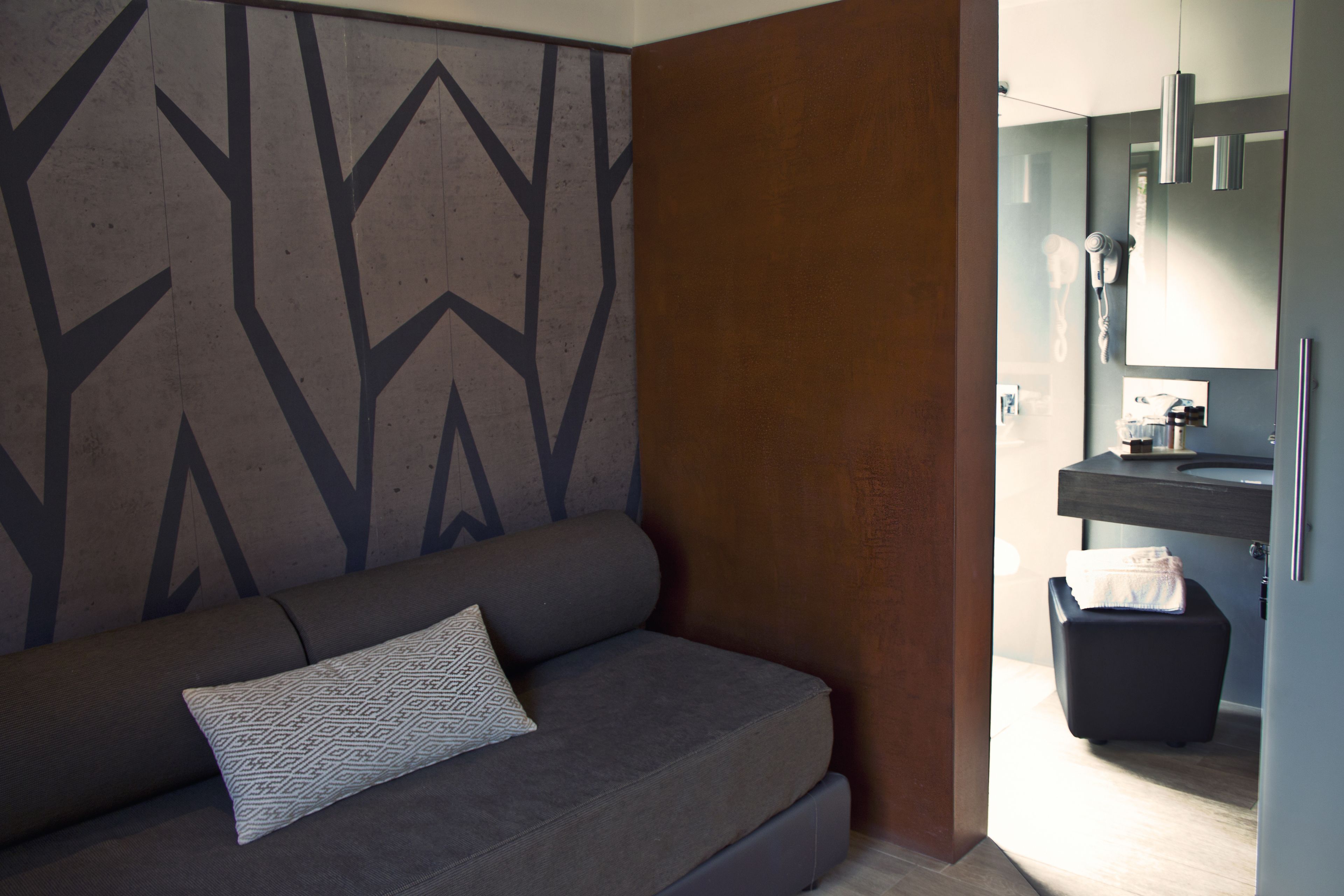 Una sala de estar moderna con un sofá gris y almohadones estampados, una pared de acento de madera y vistas a un baño con lavamanos y toallero.