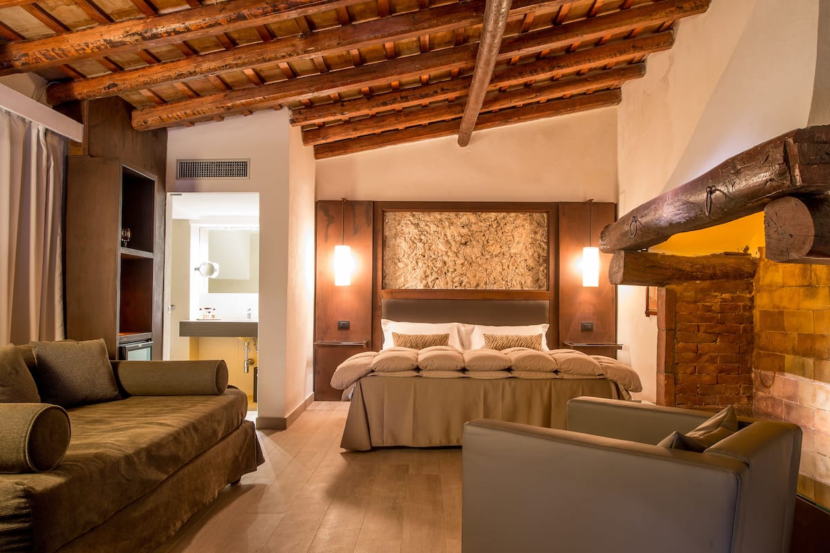 A bedroom with a wooden ceiling, a bed with multiple pillows, a sofa, and a brick wall.