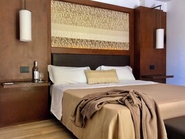 A hotel room with a large bed, a headboard with a decorative panel, a bedside table with a bottle and glass, and two wall-mounted lamps.