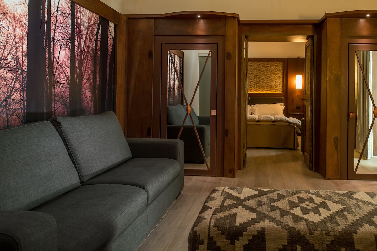 A modern hotel room with a grey sofa, a patterned rug, and a large window with a forest view.