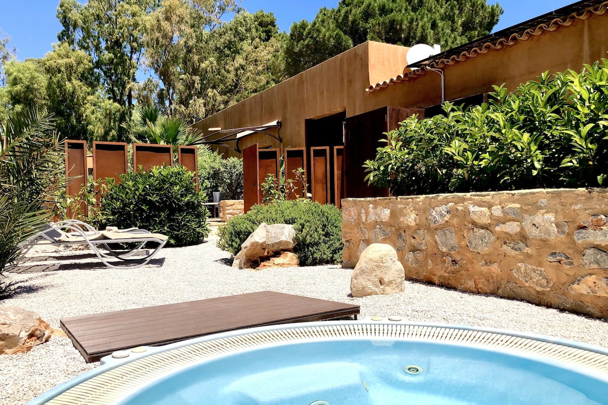 A swimming pool with a wooden deck, surrounded by a stone wall, and a building with wooden doors and a tiled roof.