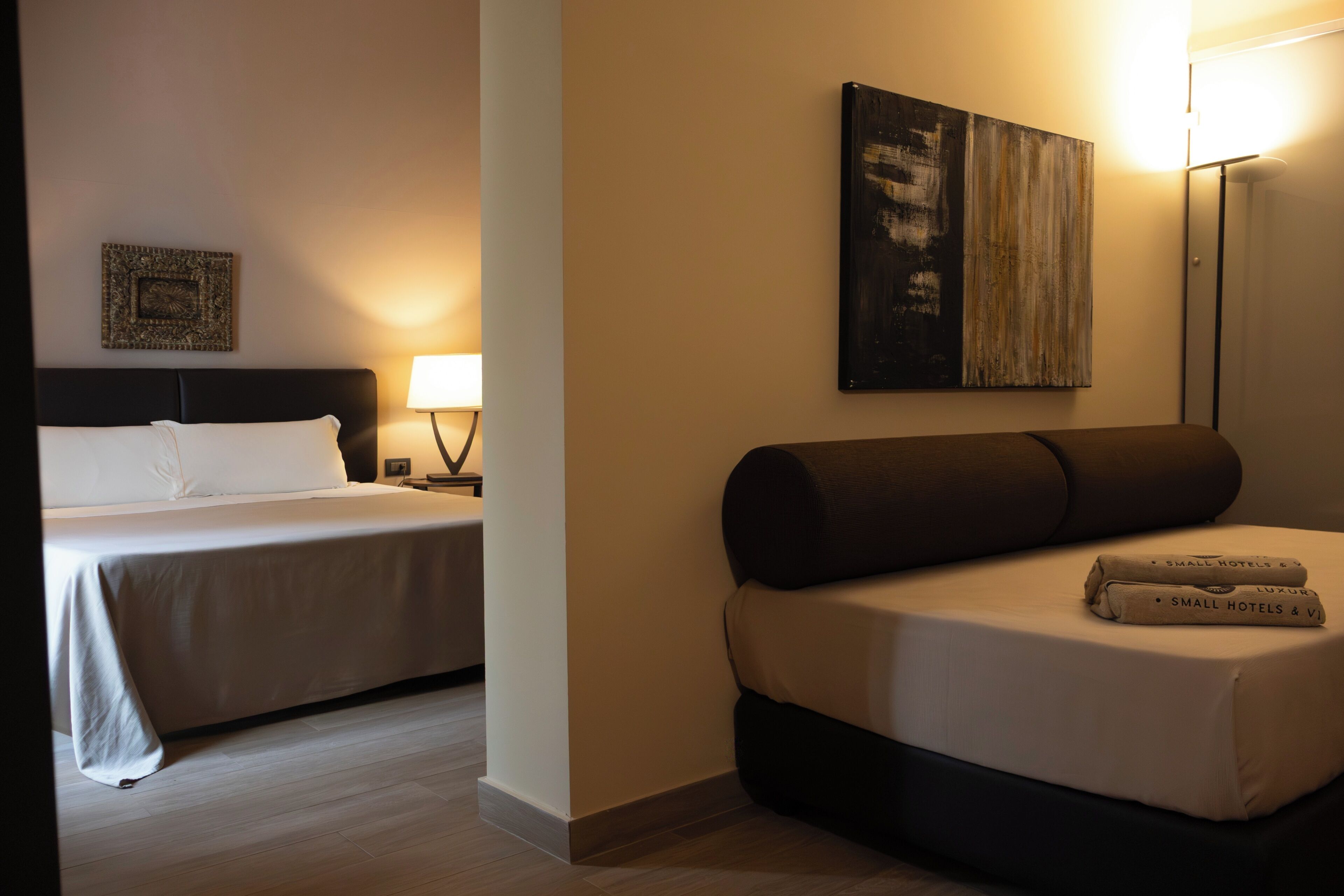 A hotel room with a bed, a sofa, a wall-mounted painting, and a nightstand with a lamp.