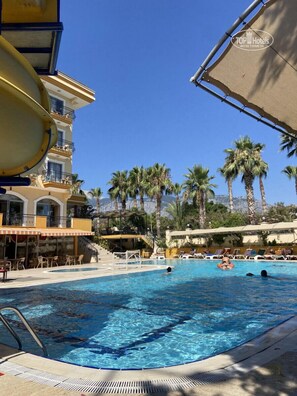 A swimming pool with lounge chairs and umbrellas, a water slide, palm trees, and a building with balconies. - Stella Hotel (Kemer)