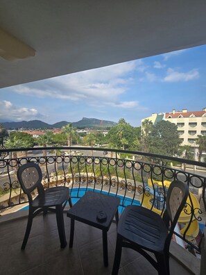 A balcony with two chairs, a small table, and a view of a swimming pool, buildings, and a hill in the distance. - Stella Hotel (Kemer)