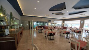 A spacious restaurant with multiple dining tables and chairs, a counter with a display, and a view of the outside. - Stella Hotel (Kemer)