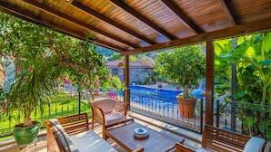 Un porche cubierto con mobiliario de madera, vista a una piscina y abundante vegetación.