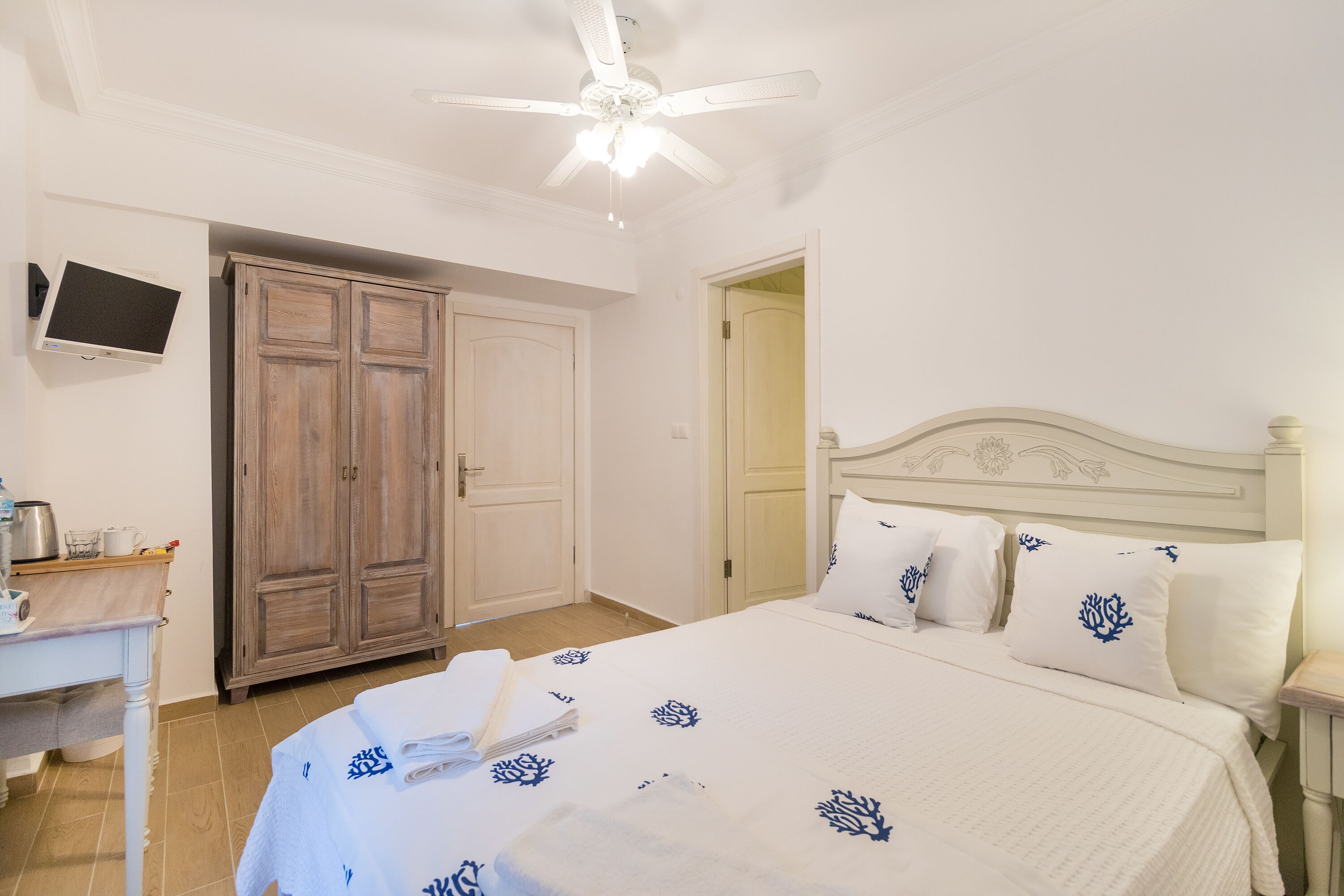 A bedroom with a bed, a wooden wardrobe, a desk with a TV, and a ceiling fan.
