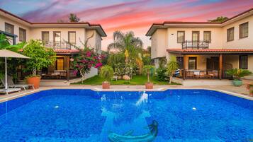 A swimming pool with a dolphin mosaic, surrounded by two-story houses with balconies, potted plants, and a clear sky.