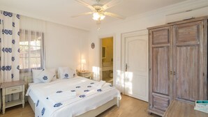 Un dormitorio con cama, mesita de noche, armario, ventilador de techo y ventana con cortinas.