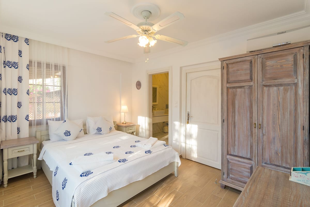 Een slaapkamer met een bed, nachtkastje, kledingkast, plafondventilator en een raam met gordijnen.