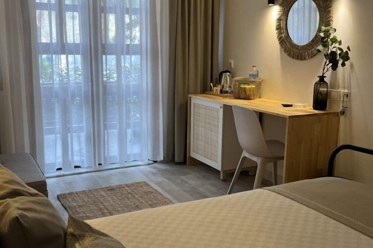 A hotel room with a bed, a desk with a chair, a radiator, a mirror, and a window with curtains.