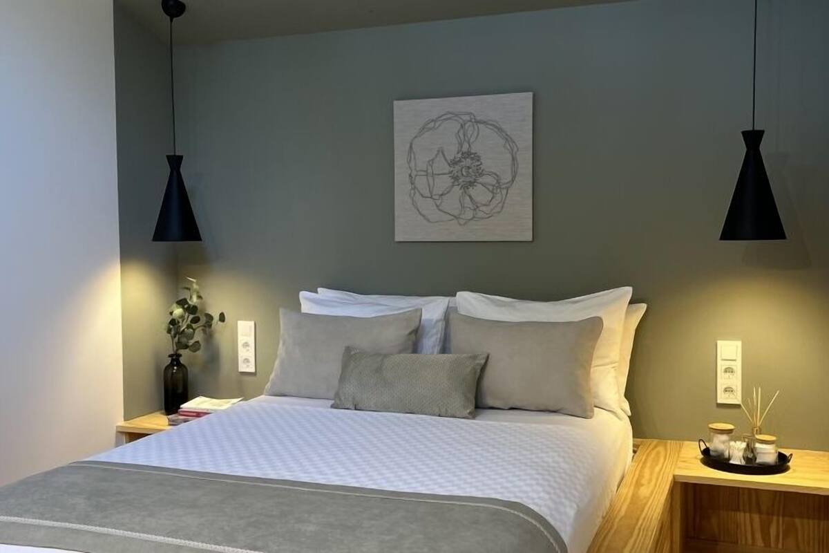 A neatly made bed with a grey blanket and pillows, a wall-mounted abstract art piece, and two black pendant lights.