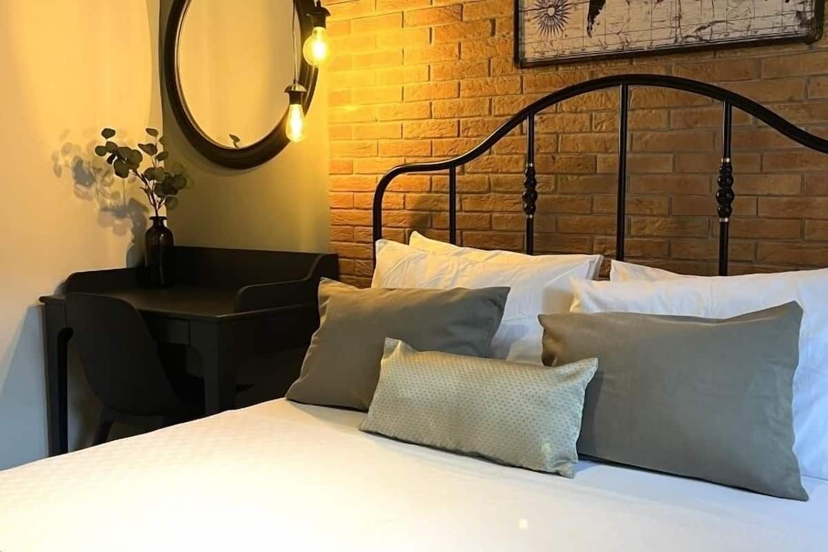 A neatly made bed with white linens and multiple pillows, a desk with a chair, a round mirror, and a wall with a brick pattern.