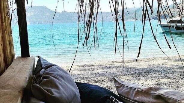 A beach view with a wooden bench, a hammock, and a boat in the water. - Selimhan Hotel - Selimiye (Marmaris)