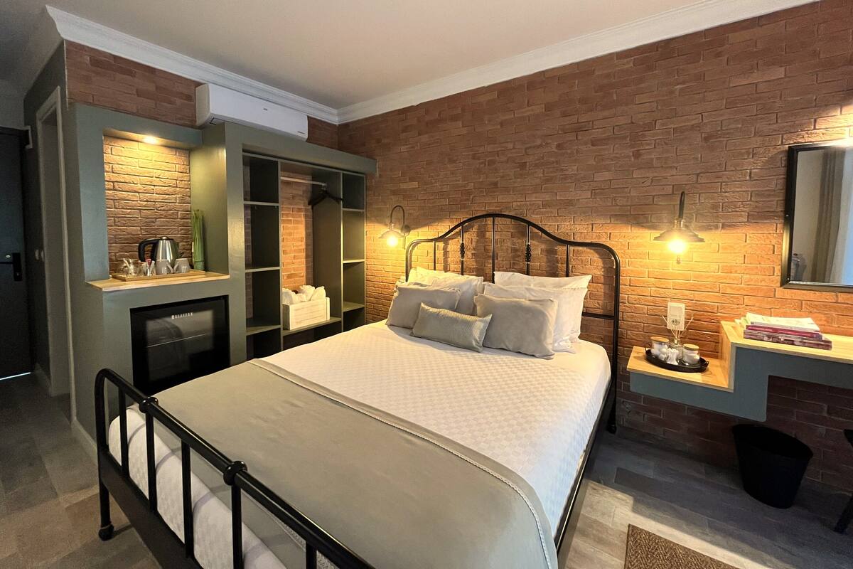 A bedroom with a brick wall, a bed with pillows, a bedside table, a mirror, and a wardrobe with shelves.