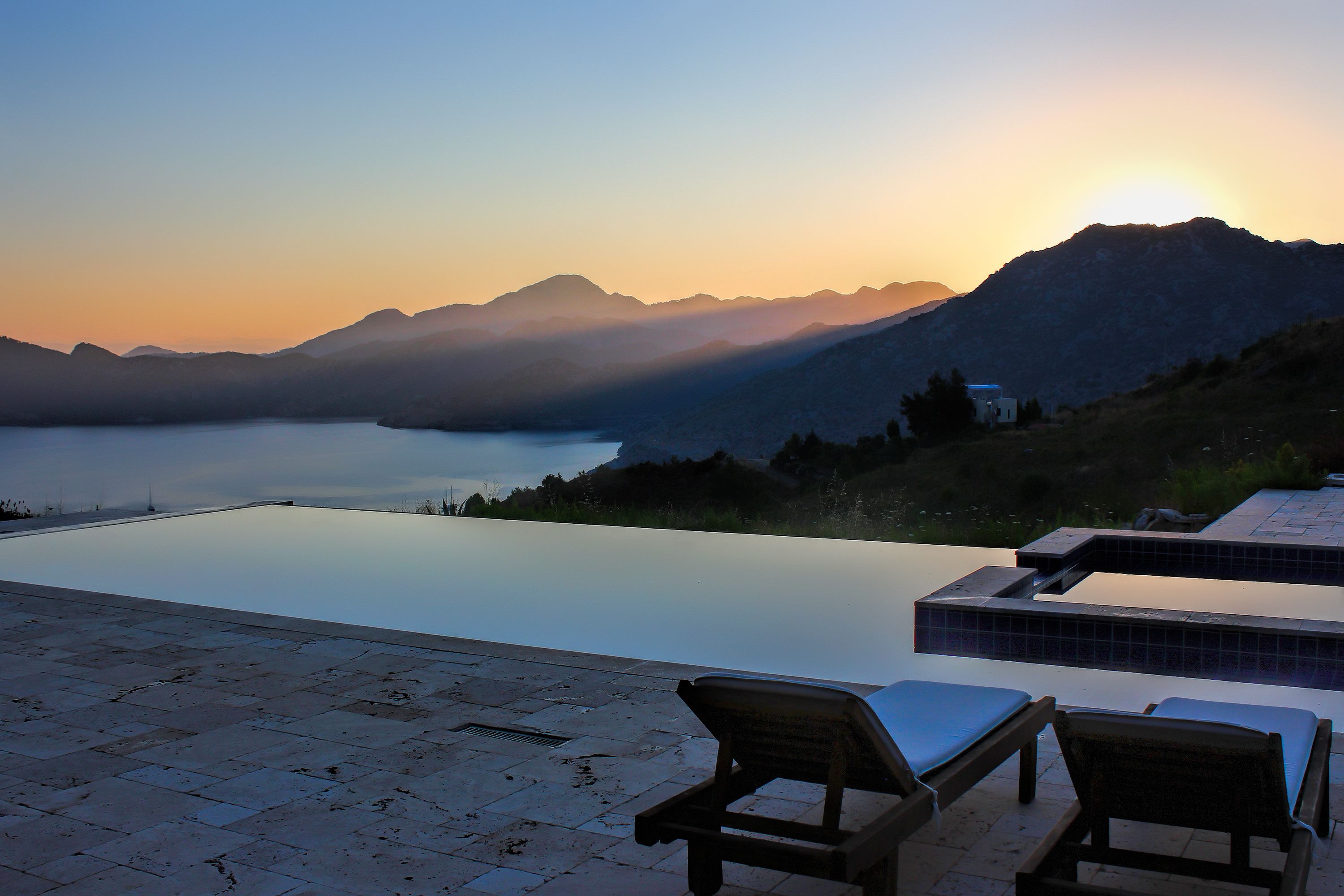 An infinity pool with two wooden lounge chairs, overlooking a lake and mountains during sunset.