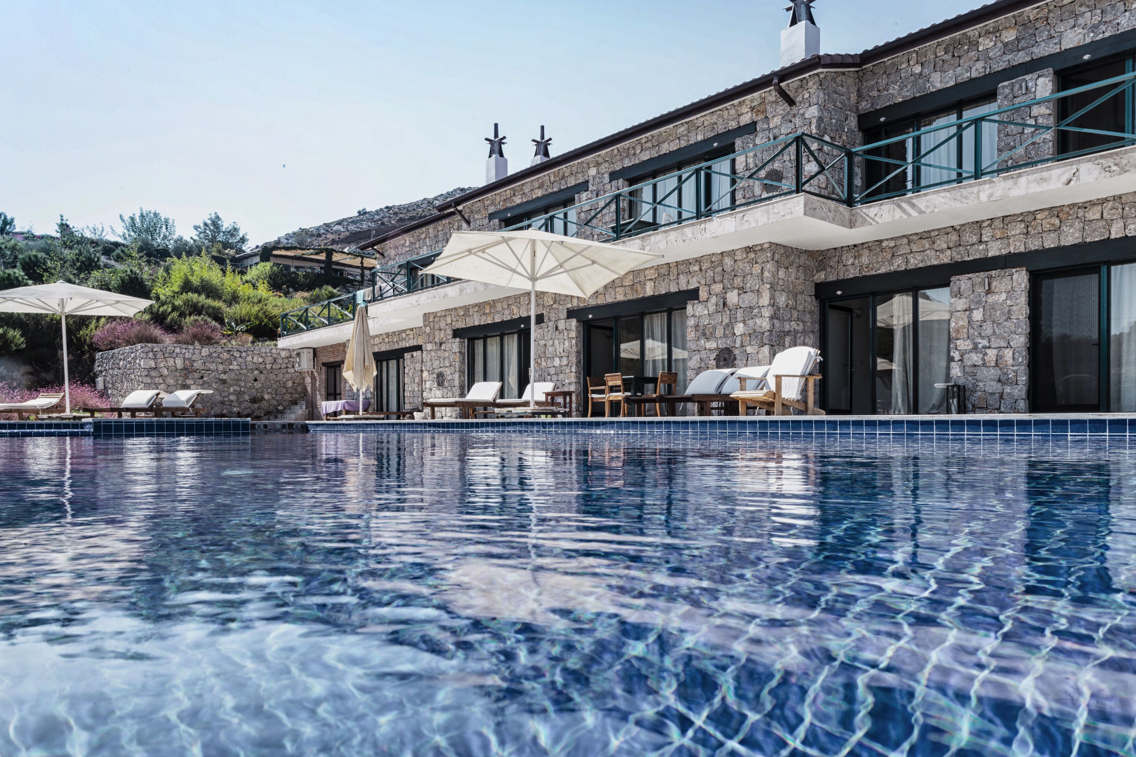 A swimming pool with lounge chairs and umbrellas, a stone building with large windows and a balcony, and a clear sky.