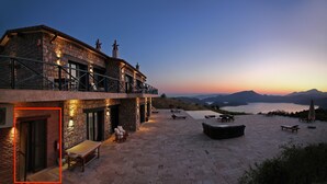 Un bâtiment en pierre doté d’un balcon offrant une vue panoramique sur un lac et des montagnes au coucher du soleil.