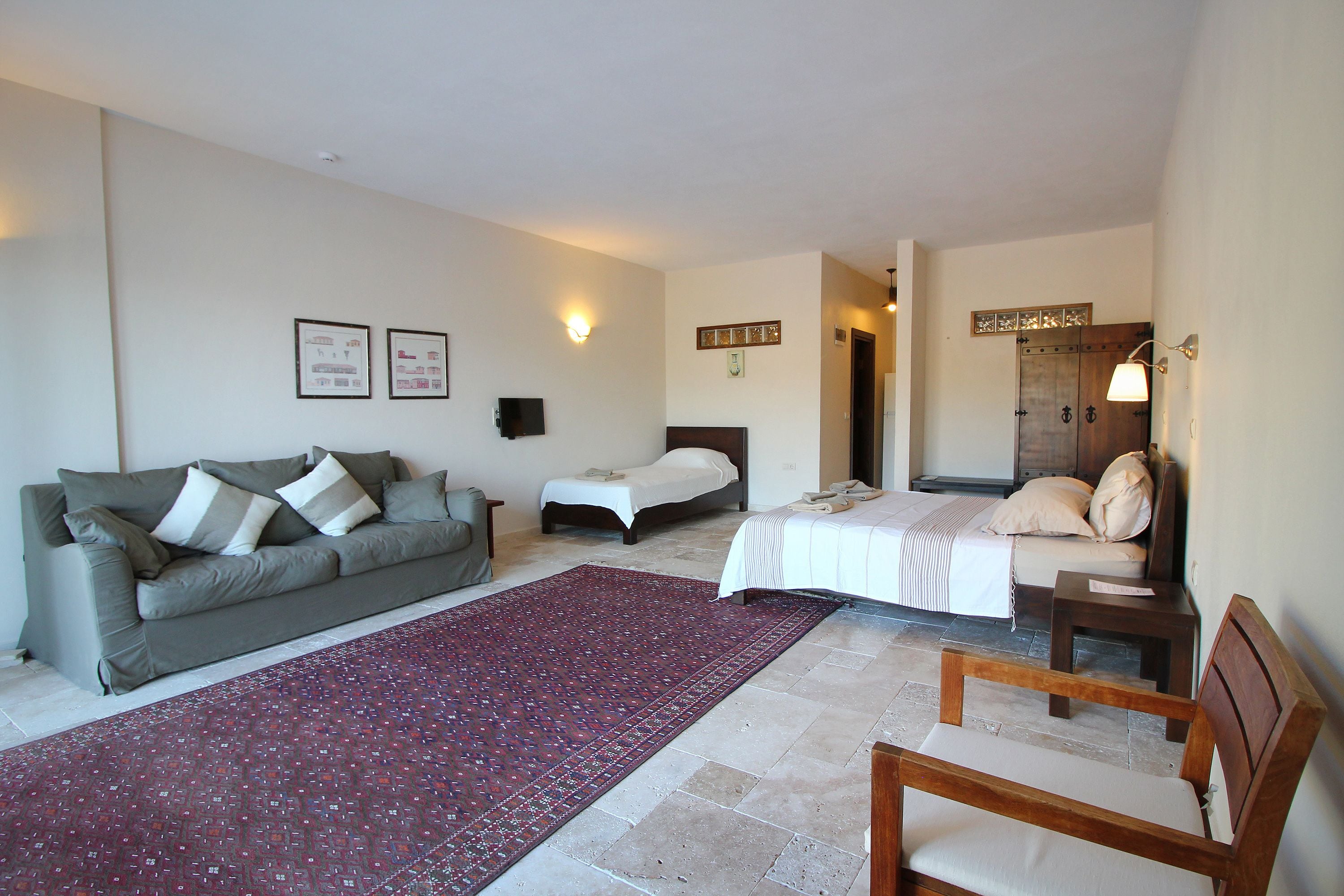A hotel room with a sofa, two beds, a wooden chair, a red patterned rug, and wall-mounted lights.