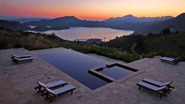 Une piscine entourée de chaises longues, avec des montagnes et un lac en toile de fond, le tout baigné par les couleurs du coucher de soleil.
