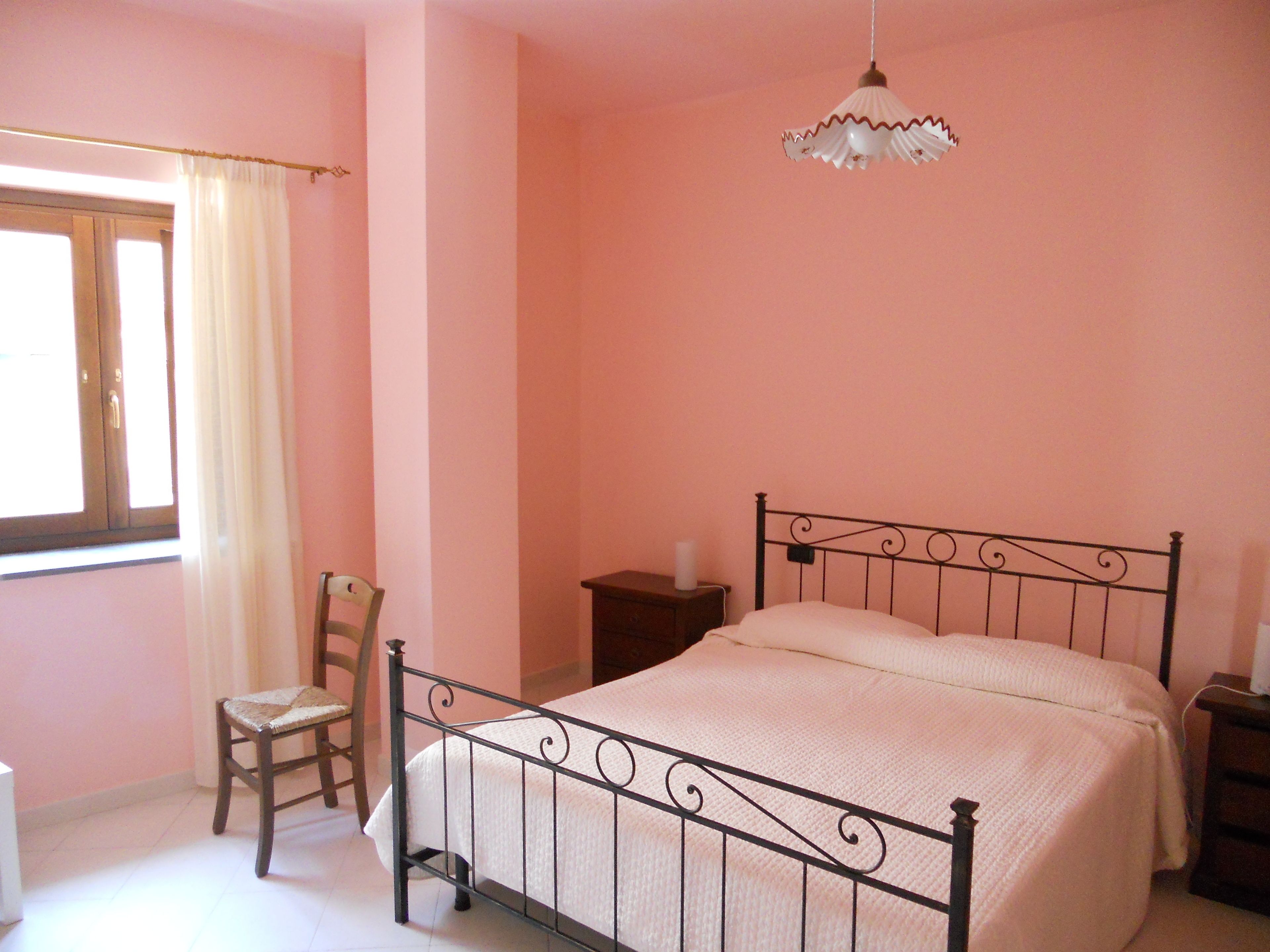 Quarto com cama, cadeira, mesa de cabeceira, luminária e janela com cortinas.