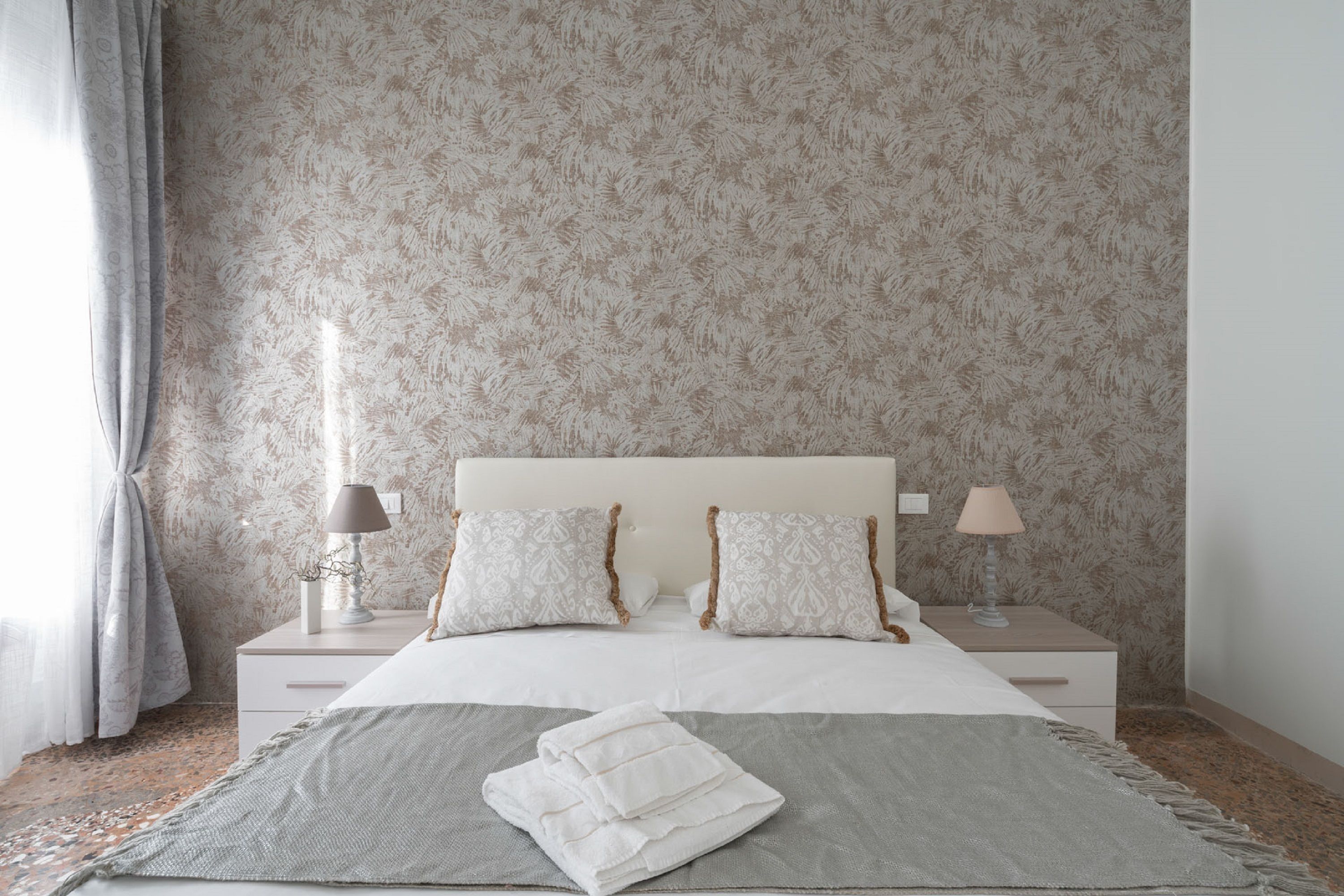 A neatly arranged bedroom with a patterned wallpaper, a bed with two pillows, two bedside tables with lamps, and a folded towel on the bed.