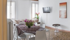 A dining area with a table set for two, a potted plant with flowers, a television on the wall, and a painting of a sunset. - Dorsoduro Charme Apartments by Wonderful Italy (Venice)