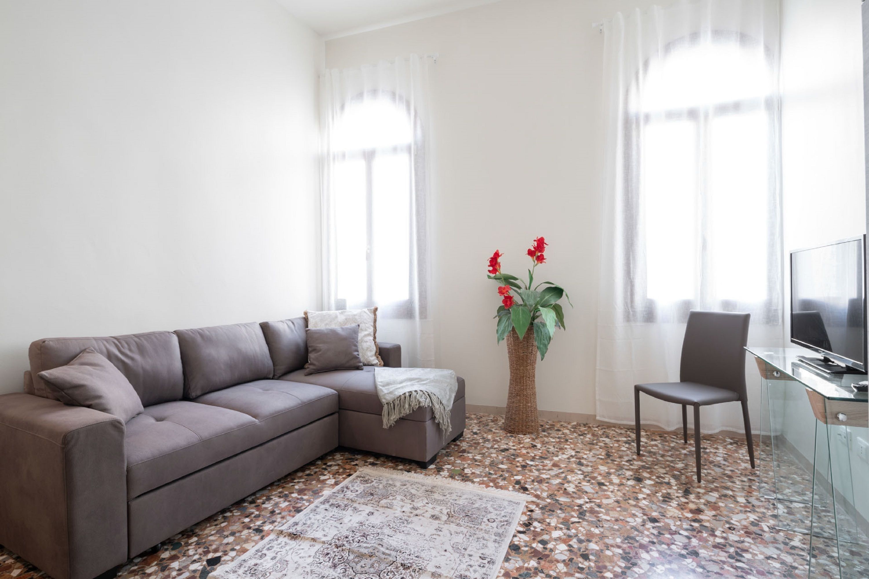 Ein modernes Wohnzimmer mit grauem Sofa, einem kleinen Tisch, einem Sessel, einem Fernseher und einer Zierpflanze.