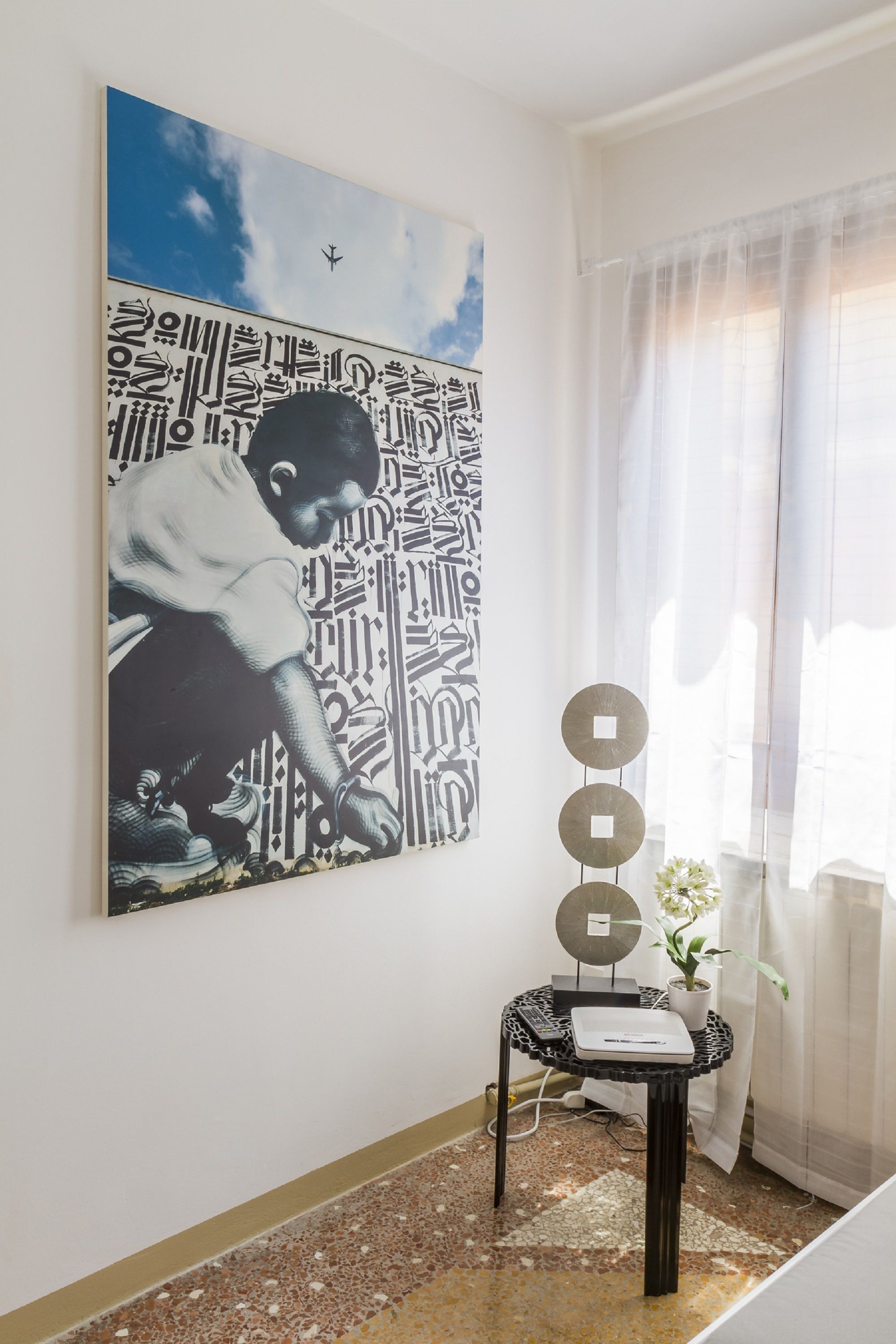 A room with a large abstract painting, a small table with decorative items, and a window with sheer curtains.