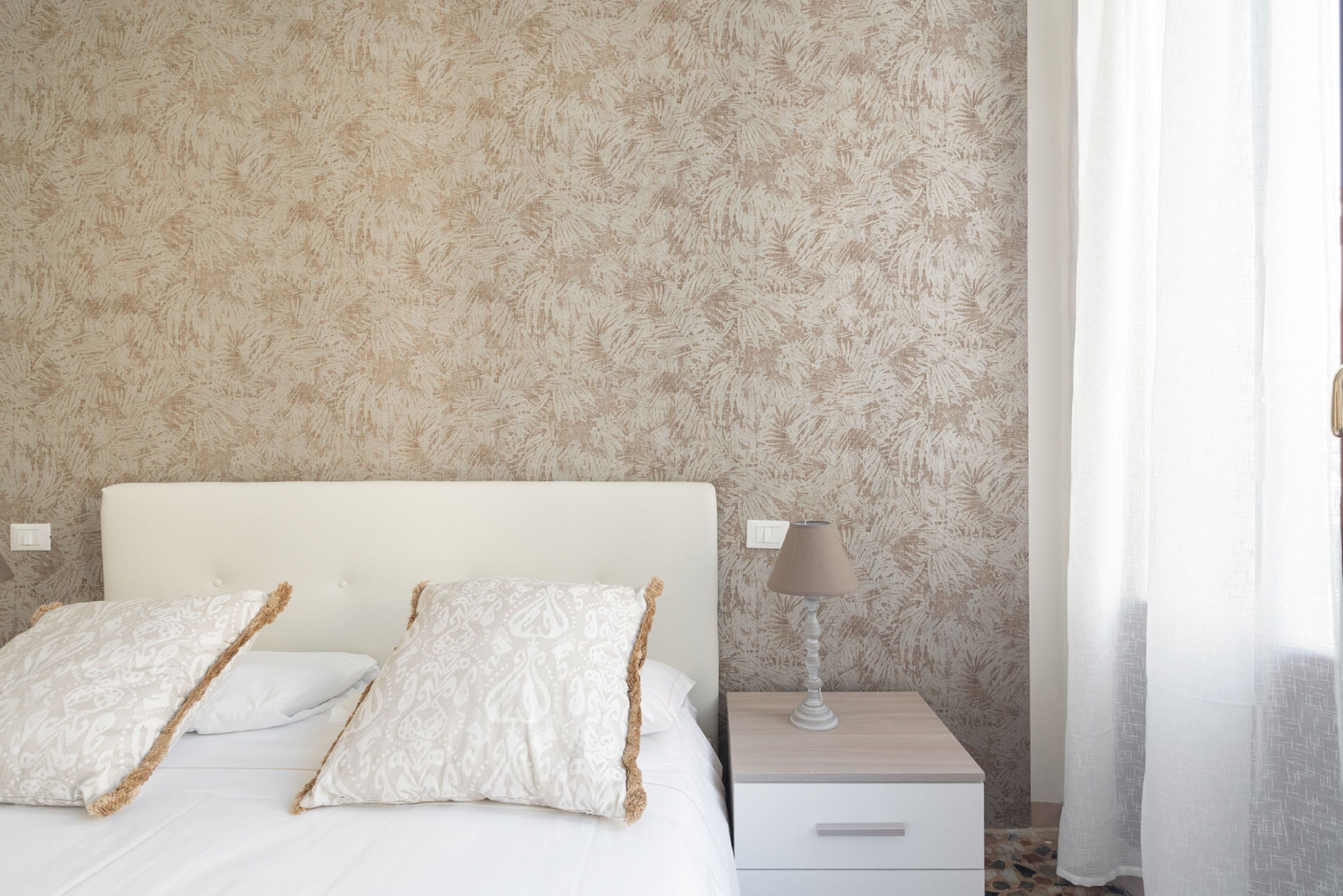 Ein ordentlich bezogenes Bett mit weißer Bettwäsche und gemusterten Kissen, ein Nachttisch mit Lampe und eine Wand mit blattmusterbehangtem Wandpapier.