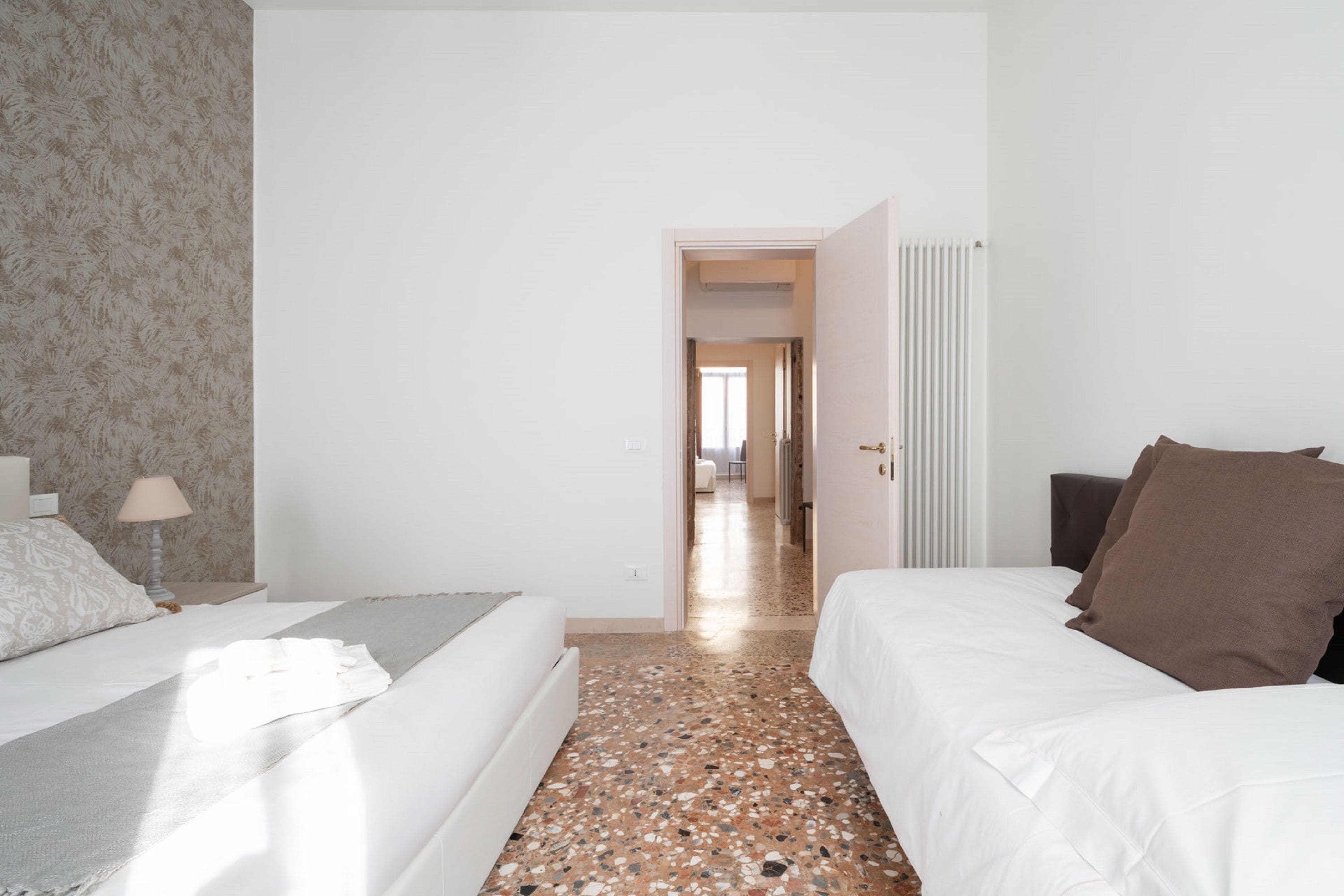 A hotel room with a bed, a nightstand, a lamp, and a door leading to another room.