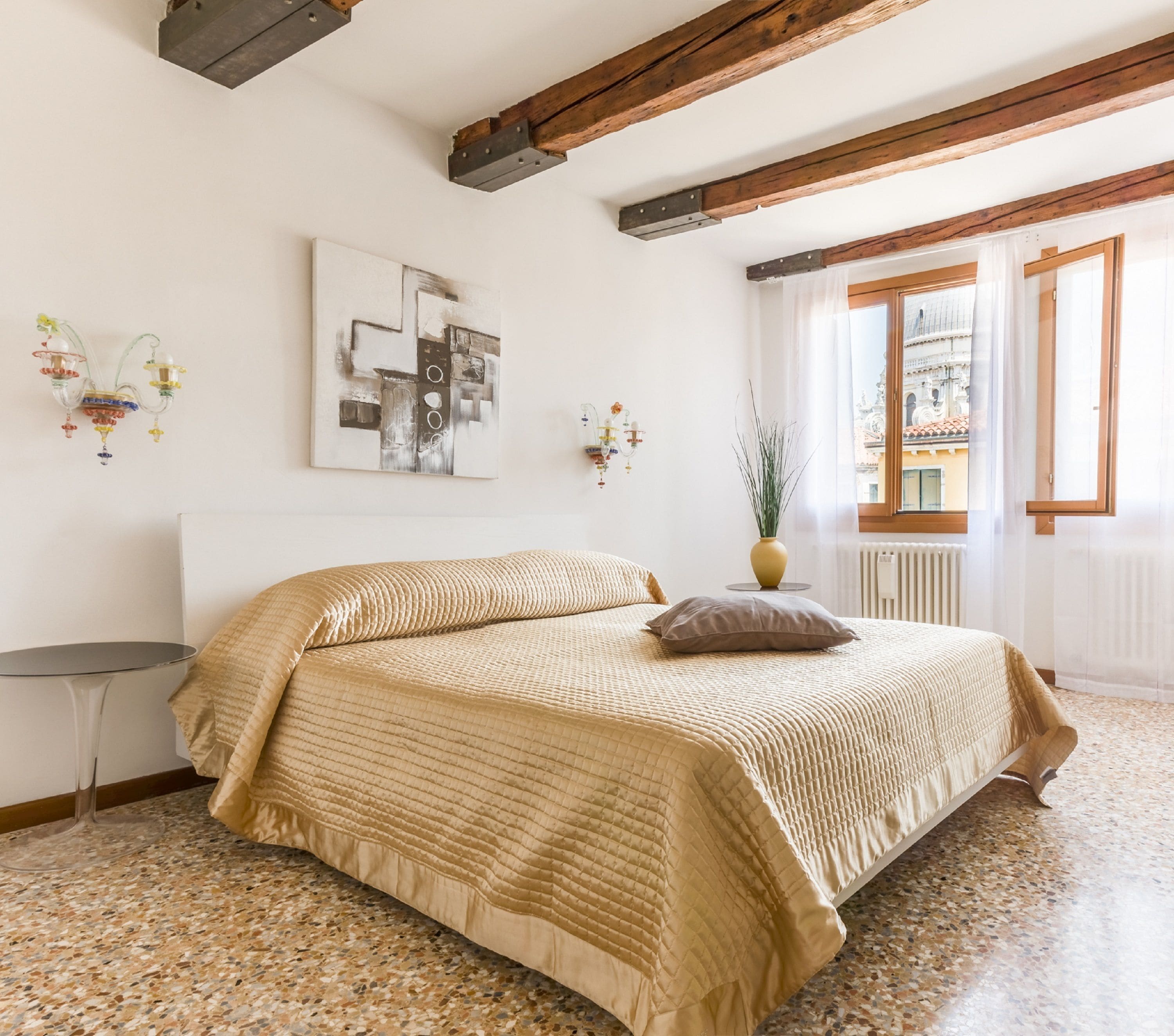 Une chambre à coucher avec un lit, une petite table, un vase avec des plantes et une fenêtre donnant sur des immeubles.