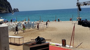Ein Strand mit weißen Liegestühlen und Sonnenschirmen, eine Bühne mit Lautsprechern und eine felsige Klippe im Hintergrund.