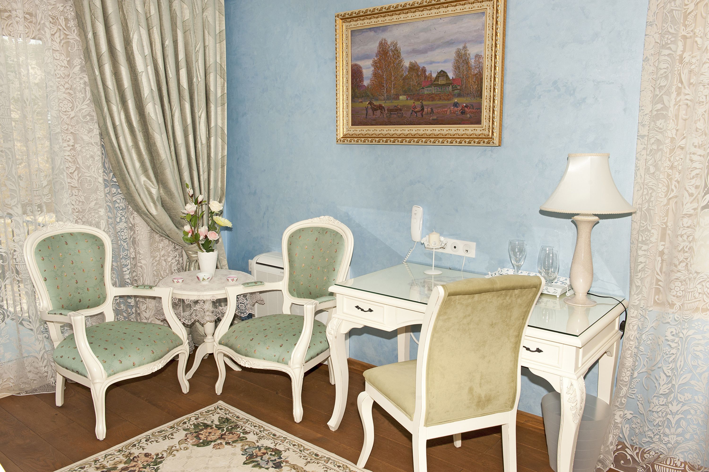 A room with a blue wall, a framed painting, a white desk, two green upholstered chairs, a white lamp, and a floral rug.