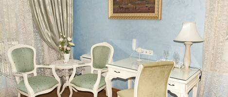 A room with a blue wall, a framed painting, a white desk, two green upholstered chairs, a white lamp, and a floral rug.