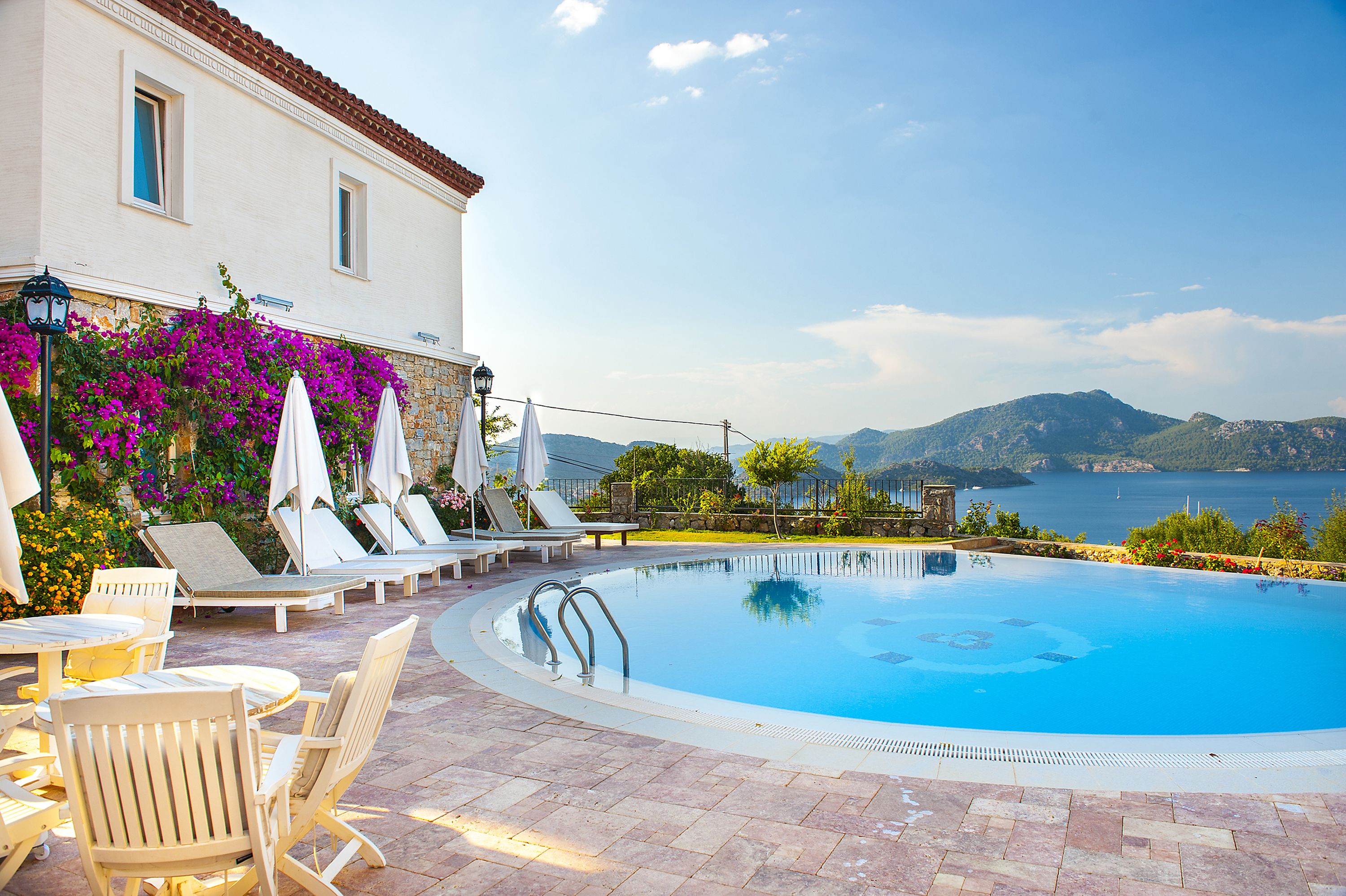 A swimming pool with lounge chairs and umbrellas, a white building with a red roof, and a view of the sea and mountains.