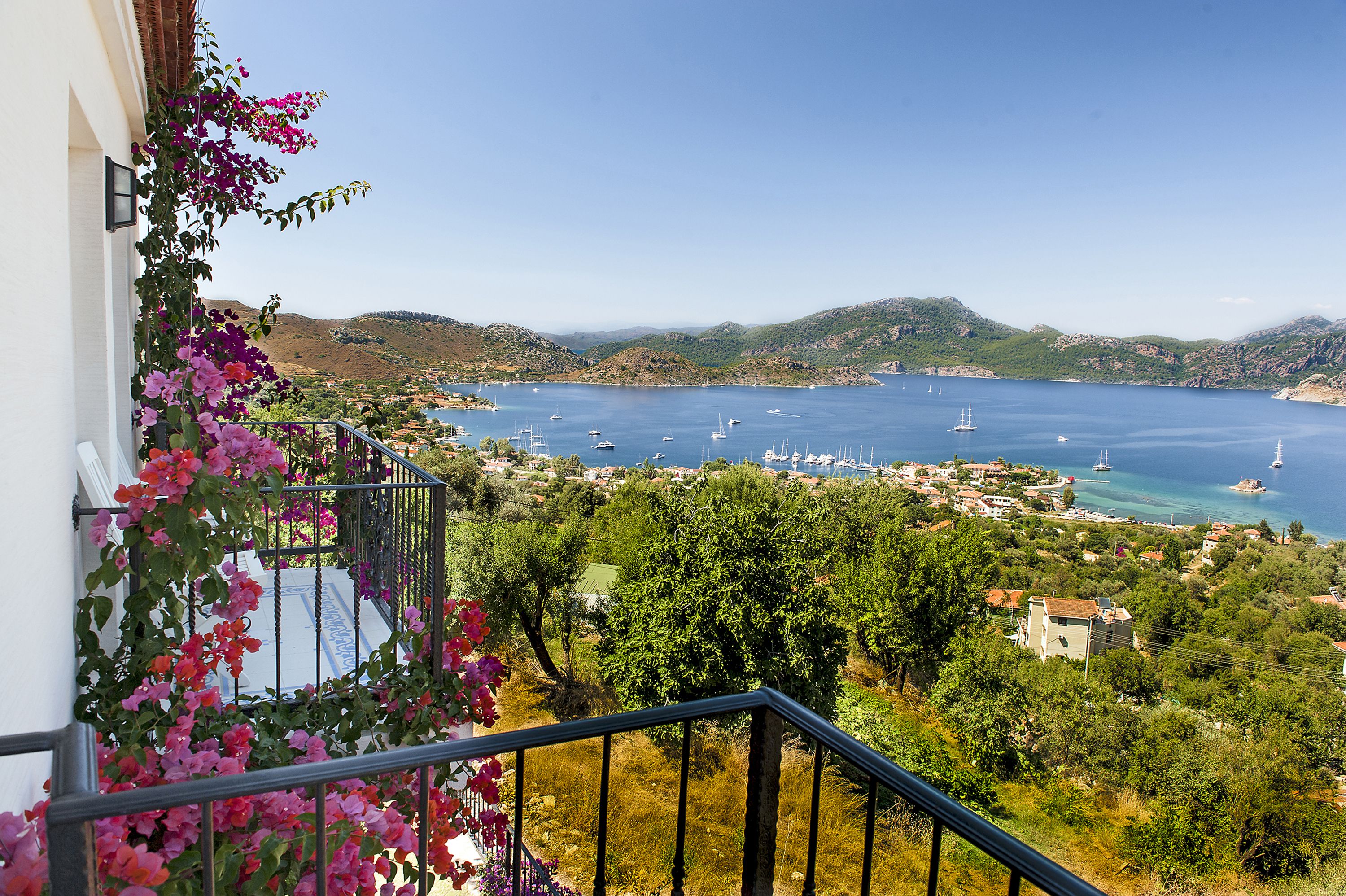 A balcony with a view of a coastal town, boats on the water, and surrounding greenery.