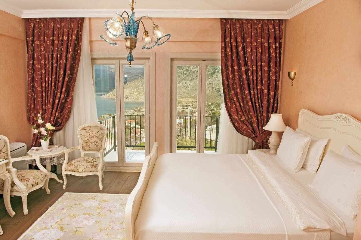 Un dormitorio con una cama grande, una araña, cortinas, una silla, una mesa y vistas al exterior.