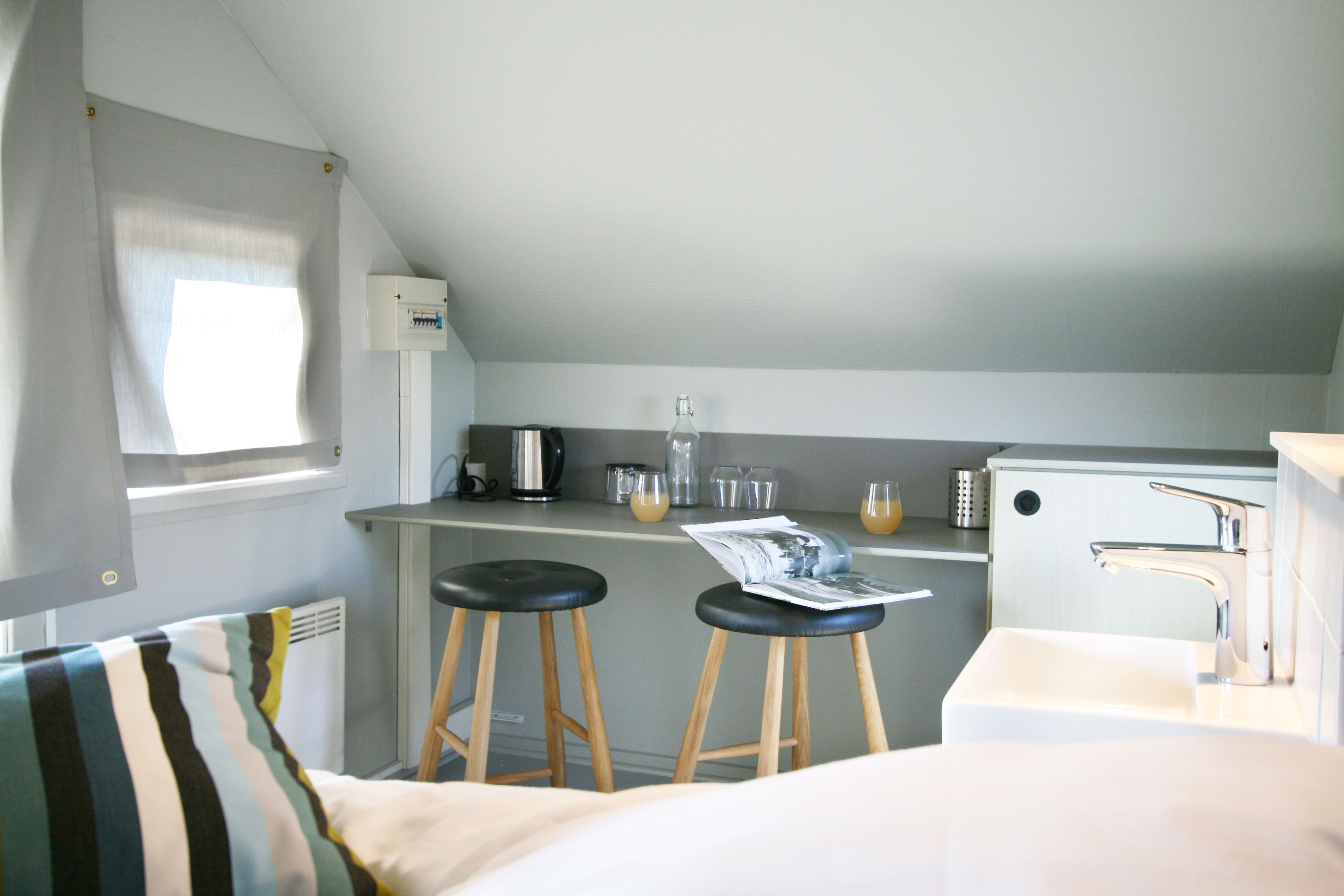 Una kitchenette compacta con un lavamanos, dos taburetes, una ventana con cortinas y un refrigerador pequeño.
