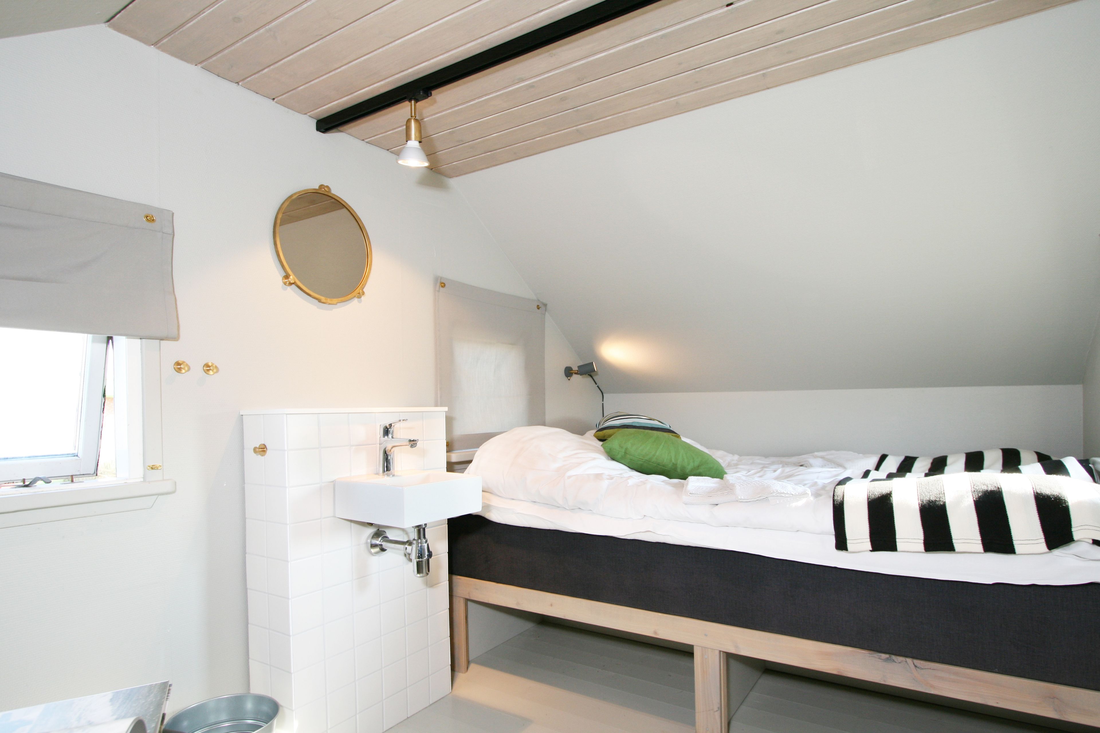 A bedroom with a wooden bed frame, a white bedside table, a mirror, and a window with a white blind.