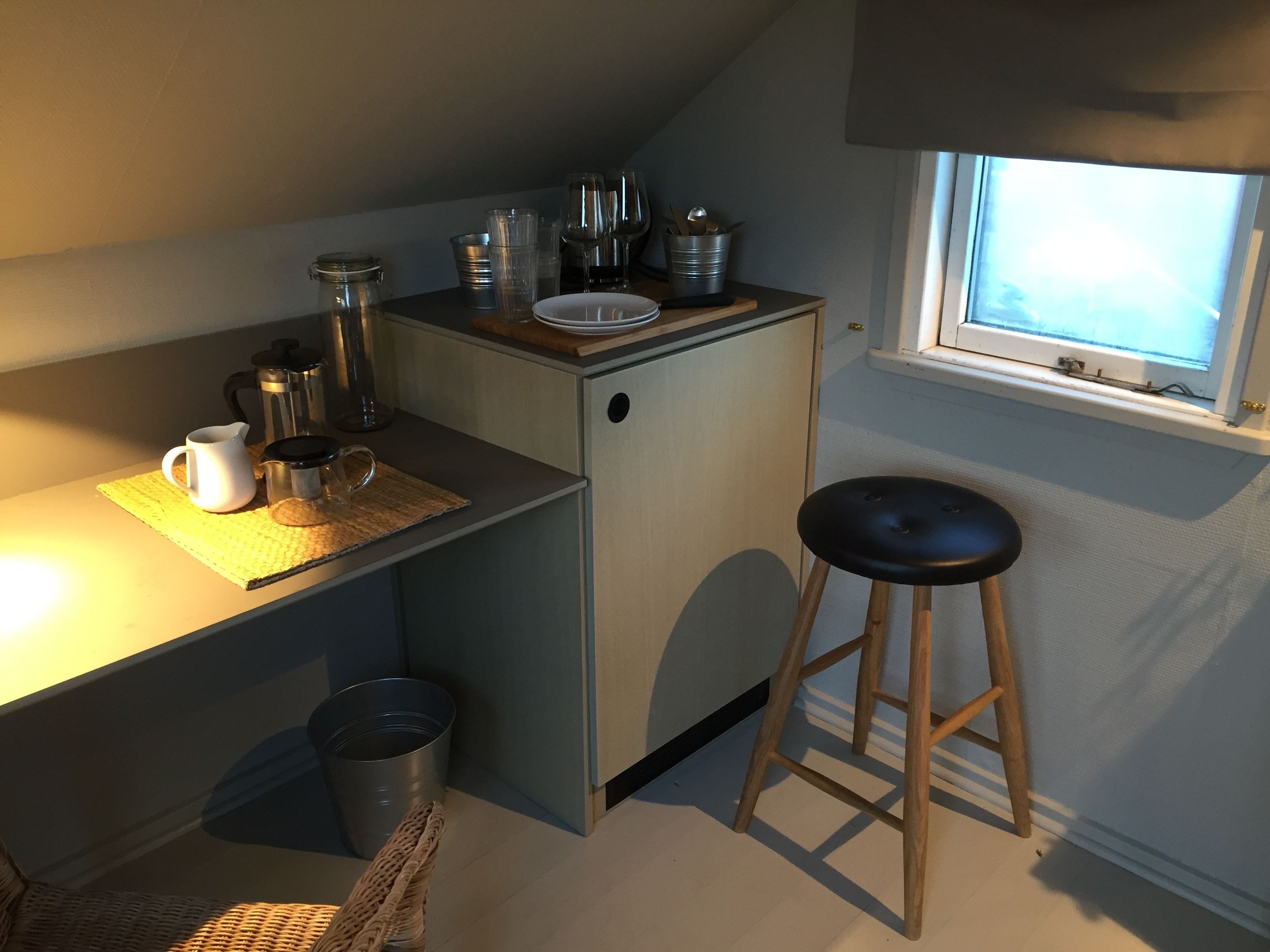 A compact kitchenette with a small refrigerator, a coffee maker, and a sink. There is a stool and a window providing natural light.