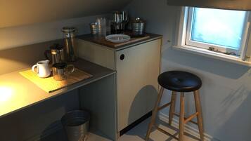 Una kitchenette compacta con un refrigerador pequeño, cafetera y pileta. Hay un taburete y un ventanal que proporciona luz natural.