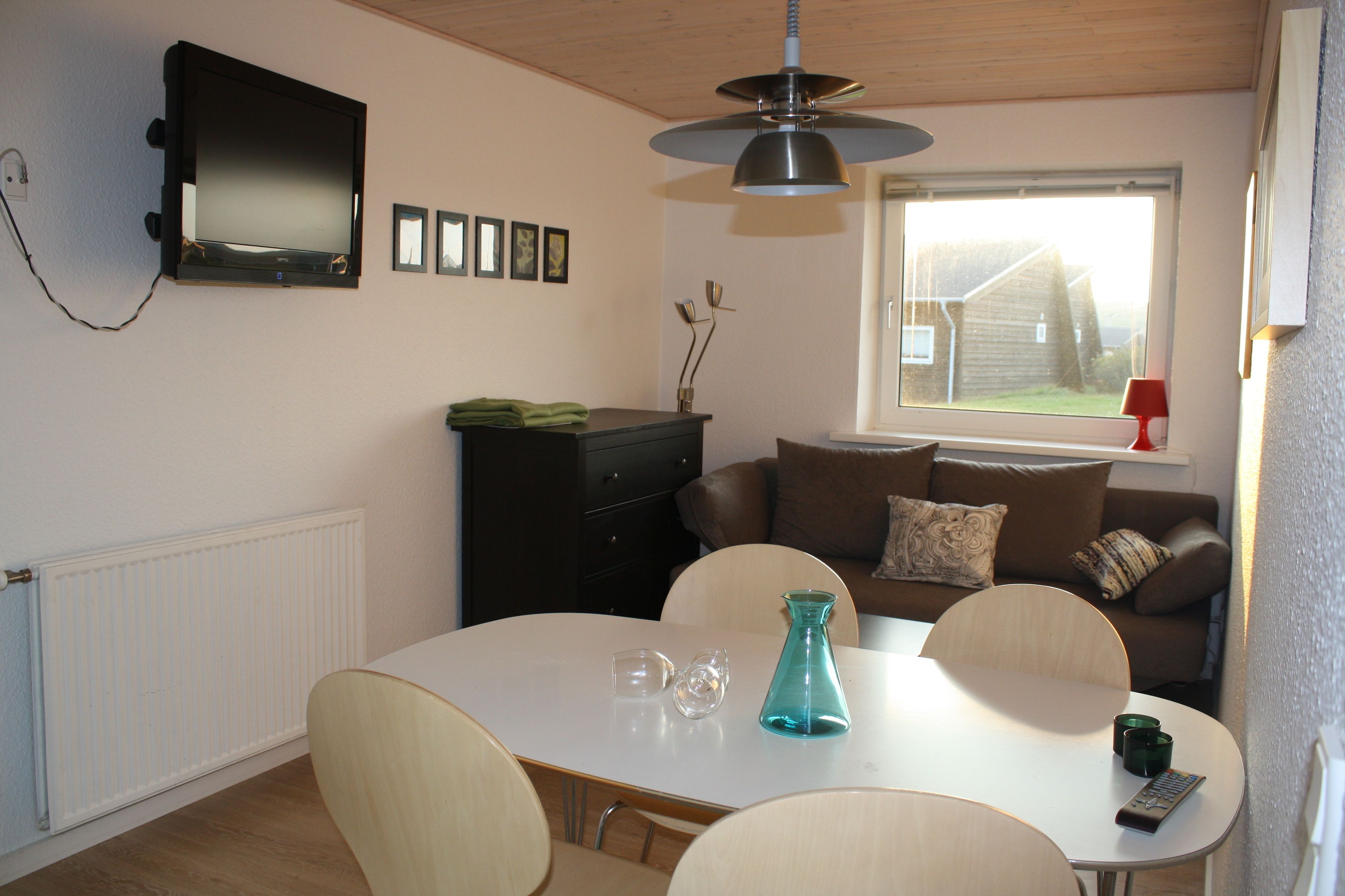 Una habitación con una mesa de comedor, sillas, un televisor montado en la pared, un ventanal con vistas a una casa y un sofá con cojines.