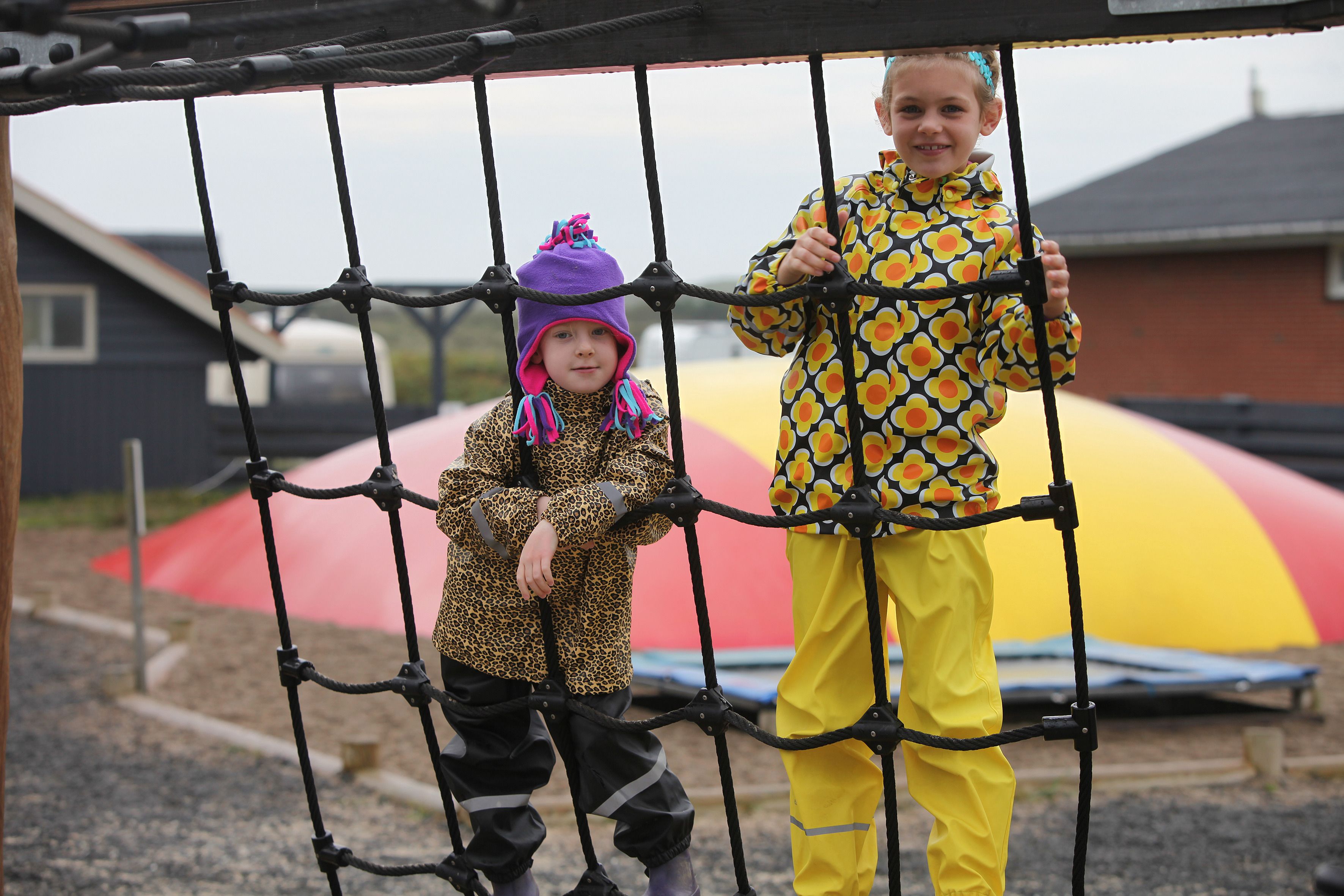 Dos niños jugando en un puente de cuerda con un tobogán inflable de colores al fondo.
