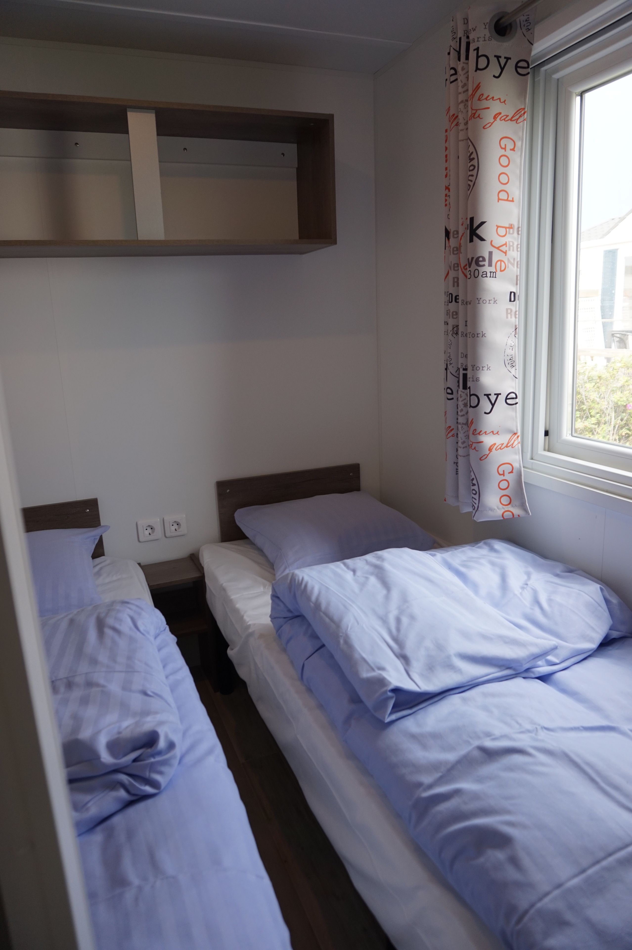 A small, single-room accommodation with two single beds, a wooden shelf above, and a window with a view.