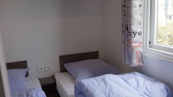 A small, single-room accommodation with two single beds, a wooden shelf above, and a window with a view.