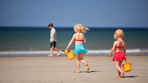 Eine Strandansicht mit zwei Kindern, die mit Eimern spielen, einem klaren Himmel und ruhiger See.